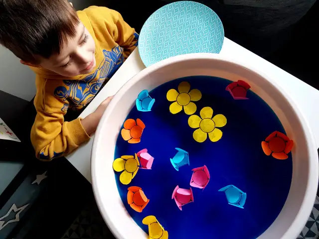 Chłopiec w żółtej bluzie obserwuje kolorowe papierowe kwiatki unoszące się na wodzie w misce.