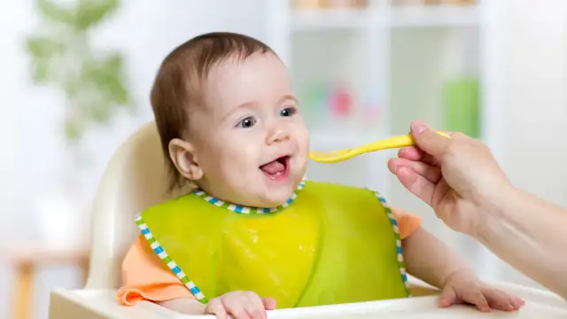Uśmiechnięte dziecko w śliniaczku dostaje łyżeczkę jedzenia dla niemowlaka.