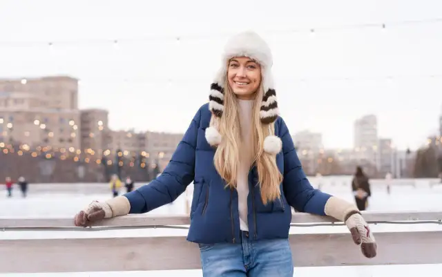 Uśmiechnięta blondynka w granatowej kurtce i białej, futrzanej czapce z pomponami. Idealny zestaw na lodowisko.