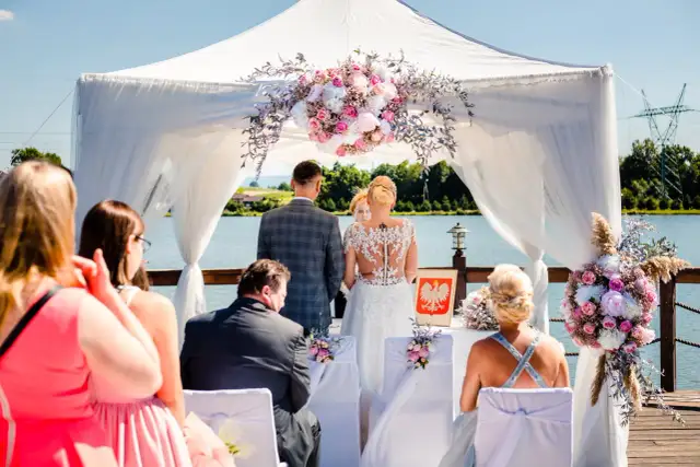 Ślub plenerowy nad jeziorem, para młoda pod baldachimem z kwiatami, goście.