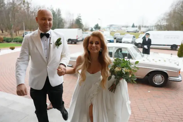 Szczęśliwi Adrian i jego wybranka, świeżo po ślubie od pierwszego wejrzenia, wychodzą na zewnątrz. Ona trzyma bukiet, on ją prowadzi.