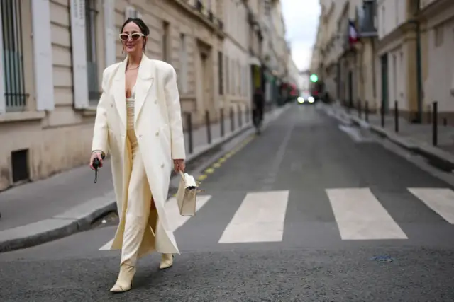 Kobieta w kremowym płaszczu i spodniach idzie ulicą, trzymając torebkę.
