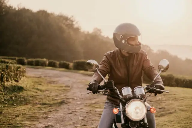 Mężczyzna w kasku i skórzanej kurtce jedzie na motocyklu po polnej drodze. Słońce zachodzi, tworząc ciepłą, złotą poświatę.