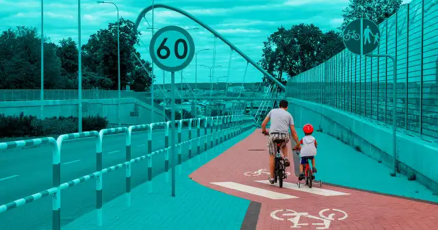 Ojciec i dziecko jadą rowerami po ścieżce rowerowej obok drogi.