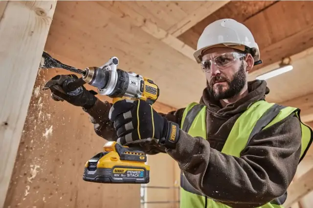 Budowlaniec w kasku i okularach wierci w drewnie wiertłem łopatkowym. Widać wyraźne symbole na wiertarce DeWalt.