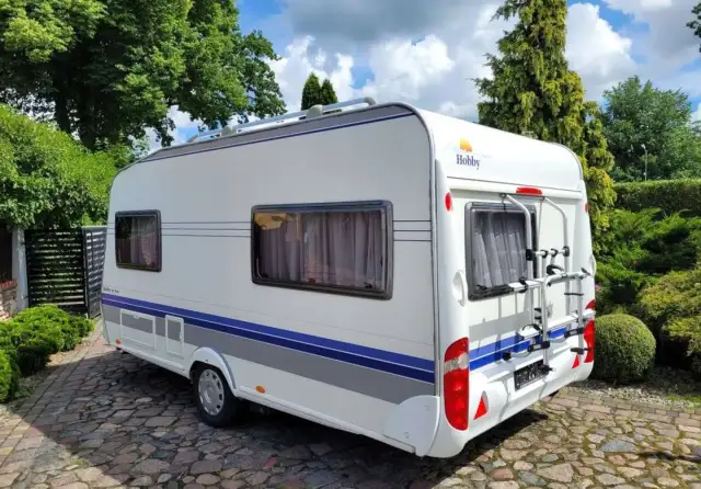 Biała przyczepa kempingowa Hobby 440 z niebieskimi pasami, zaparkowana na brukowanym podjeździe. Idealna do podróżniczego hobby.