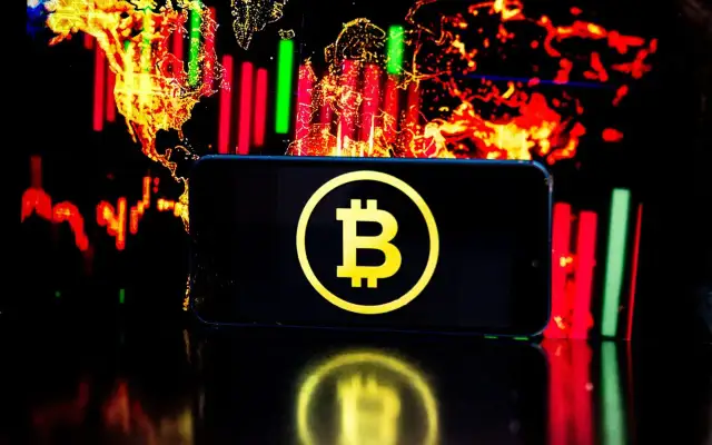 Telefon z symbolem Bitcoin na tle wykresów giełdowych. Czy warto jeszcze inwestować w bitcoin? To pytanie zadaje sobie wielu.