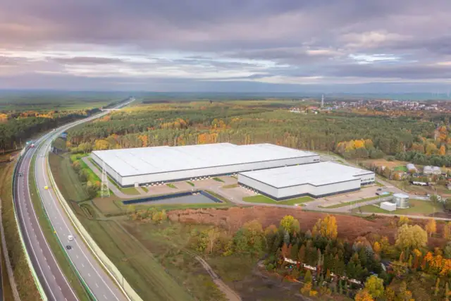 Nowoczesne, rozległe budynki firm magazynowych otoczone jesiennym lasem, w pobliżu autostrady.