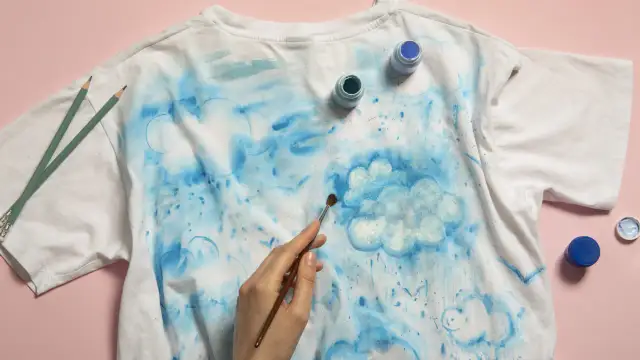 Rysowanie koszulki z chmurkami. Pędzel maluje białą chmurę na tle niebieskich rozbryzgów. Obok leżą farby i ołówki.