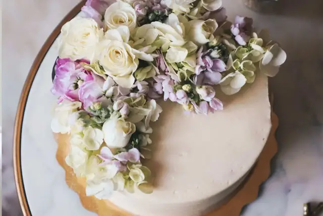 Elegancki tort z kremową polewą, ozdobiony bukietem żywych goździków, róż i hortensji w pastelowych kolorach.