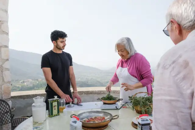 An older woman in an apron cooks greens while a young man and an older man stand nearby on a terrace with a scenic view.