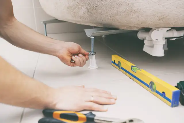 Ręka dokręca śrubę wspierającą brodzik, obok leży poziomica. To kluczowy etap, jak zamontować brodzik wysoki.