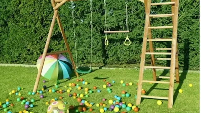 Drewniany plac zabaw ze zjeżdżalnią, huśtawką i drabinką, otoczony kolorowymi piłeczkami i parasolem.