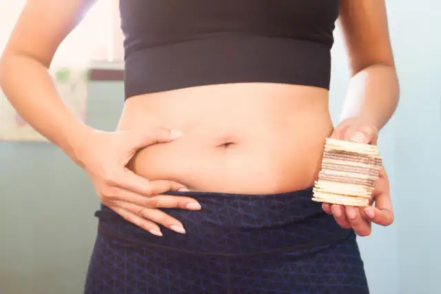 Kobieta chwyta fałd tłuszczu na brzuchu, trzymając w dłoni batonik. Czy to wyzwanie, jak schudnąć z brzucha po 40?