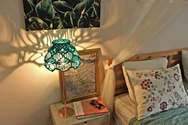 Zielona, pleciona lampa nocna rzuca cienie na ścianę. Obok łóżko z poduszką.