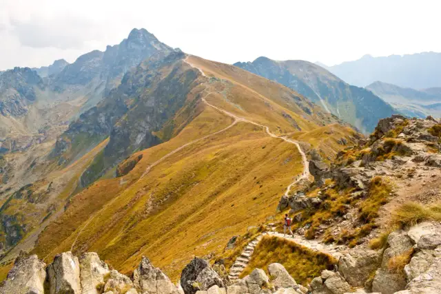 Szlak turystyczny wije się pośród skalistych zboczy, prowadząc ku majestatycznym, jesiennym szczytom. W oddali widać kolejne pasma górskie.