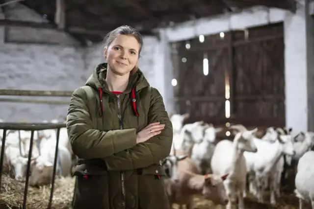 Kobieta w zielonej kurtce stoi na tle stada kóz. Jej uśmiech sugeruje, że hodowla kóz opłacalność jest dla niej satysfakcjonująca.
