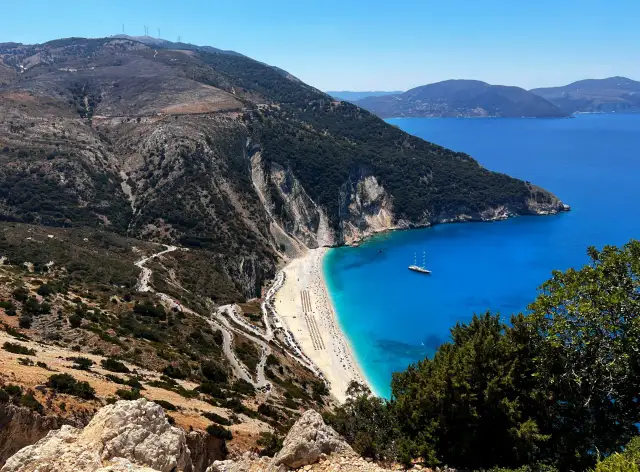 Widok na plażę Myrtos, jedno z najpiękniejszych miejsc na Kefalonii. Kręta droga prowadzi do białego piasku i turkusowej wody.