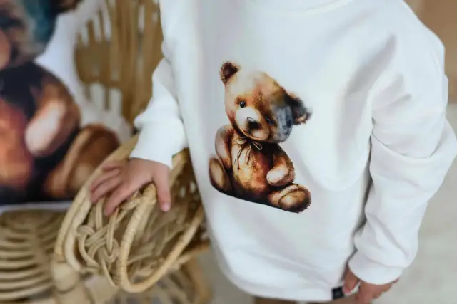 Biała bluza Moschino z uroczym misiem w stylu vintage. Dziecko trzyma się wiklinowego koszyka.