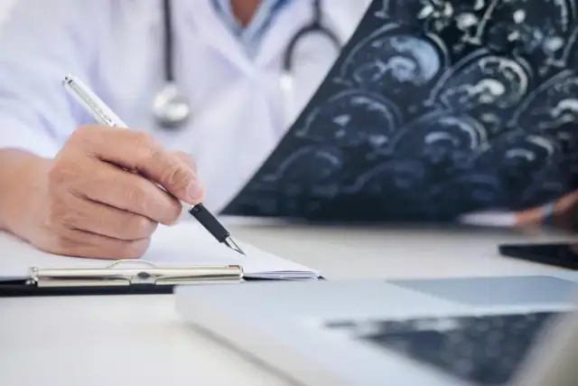 Lekarz w białym kitlu analizuje zdjęcie rezonansu magnetycznego mózgu i pisze notatki.