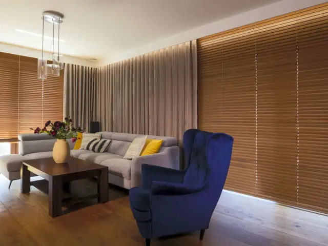 Elegancki salon z szarym narożnikiem, granatowym fotelem i drewnianymi żaluzjami oraz zasłonami w ciepłych odcieniach.