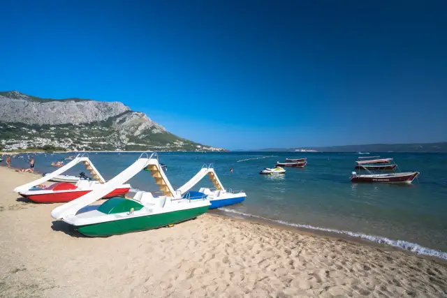 Plaża z łódkami i zjeżdżalniami, idealna na wakacje w camping Galeb. W tle góry i turkusowe morze.