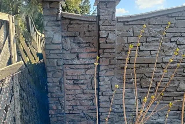 Betonowy płot z fakturą kamienia, z widocznymi słupkami i fragmentem starego płotu.