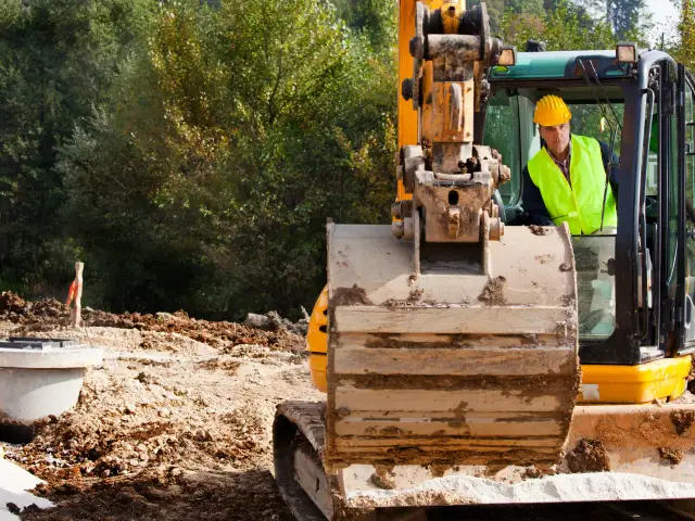 Operator koparki w żółtym kasku i kamizelce odblaskowej siedzi w kabinie maszyny na placu budowy.
