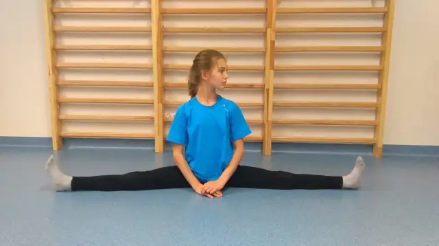 Dziewczynka w niebieskiej koszulce robi szpagat poprzeczny na niebieskiej podłodze, na tle drabinek gimnastycznych.