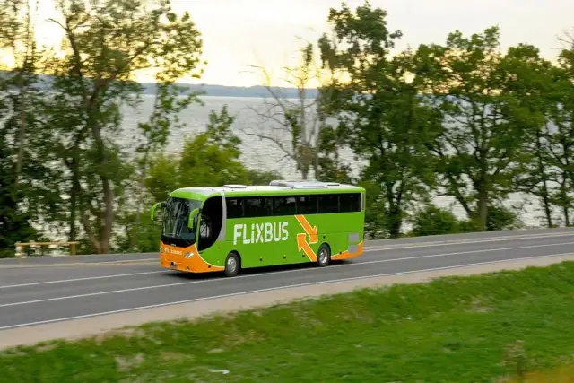 Zielony autobus Flixbus jedzie drogą przy jeziorze otoczonym drzewami.