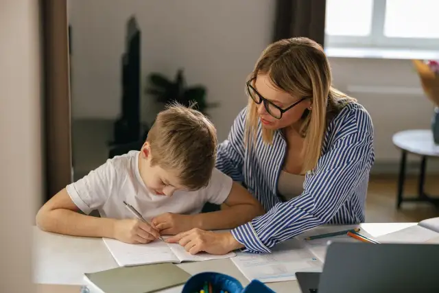 Kobieta w okularach pomaga chłopcu w nauce przy biurku.