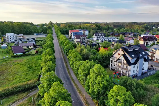 Droga w otoczeniu zieleni prowadzi do centrum Jastrzębiej Góry, gdzie widać domy i apartamentowce.