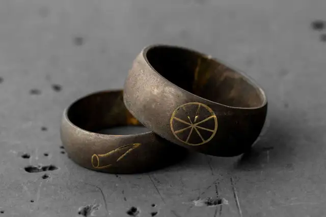 Dwie czarne obrączki męskie z złotymi symbolami: kołem z promieniami i stylizowanym rogiem.