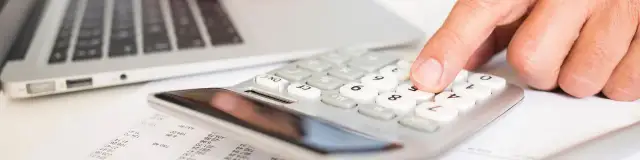 Dłoń naciska klawisze kalkulatora obok laptopa i dokumentów.