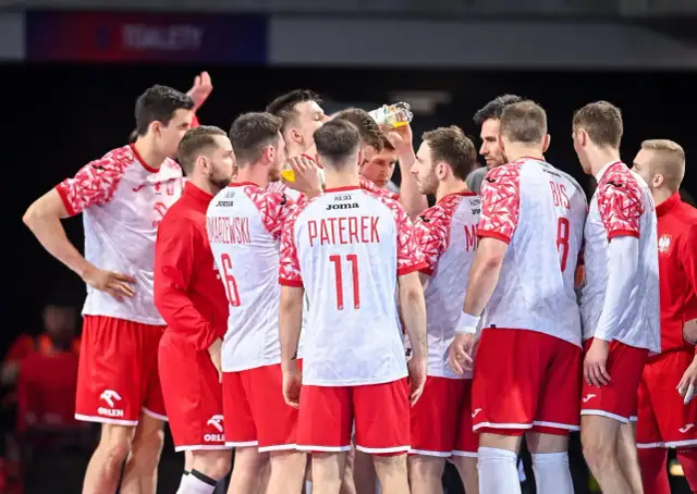 Drużyna piłkarzy ręcznych Polski w czerwono-białych strojach w skupieniu słucha trenera.