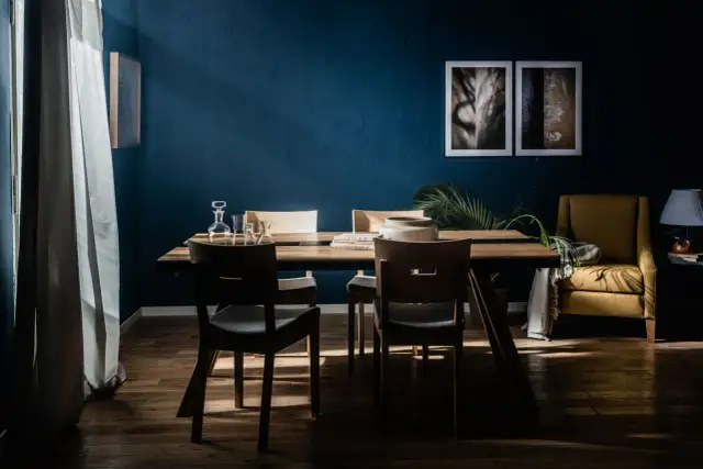 Jadalnia w głębokich odcieniach niebieskiego, z drewnianym stołem i krzesłami, żółtym fotelem i roślinnością.