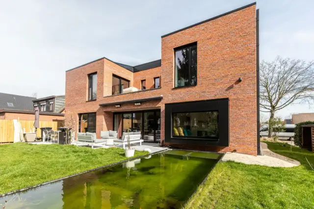 Nowoczesny dom z cegły z dużymi oknami i tarasem. W ogrodzie znajduje się zielony staw. Okiennice dodają mu charakteru.