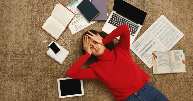 Zmęczona studentka leży na dywanie otoczona książkami, laptopem i telefonem.