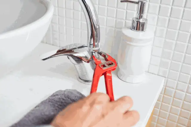 Dłoń z czerwonymi szczypcami dokręca chromowany kran umywalkowy.