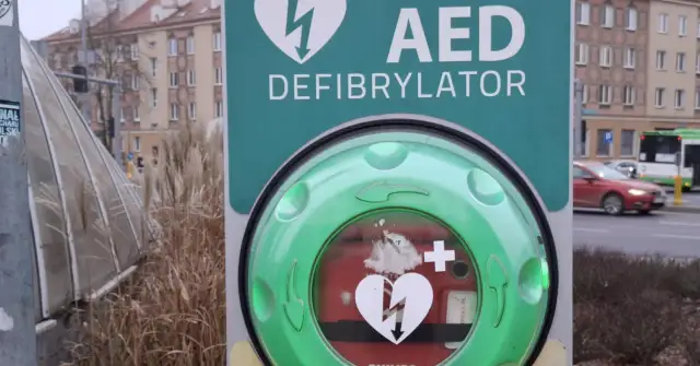 Zielony AED (defibrylator) z symbolem serca i pioruna na tle miejskim.