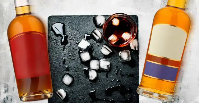 Dwie butelki, jedna z czerwoną etykietą, druga z beżową i fioletową, stoją obok szklanki z lodem i ciemnym płynem. Koniak czy whisky?