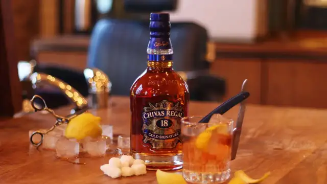 Butelka Chivas Regal 18, szklanka z drinkiem, kostki lodu, cytryna i cukier na drewnianym stole.