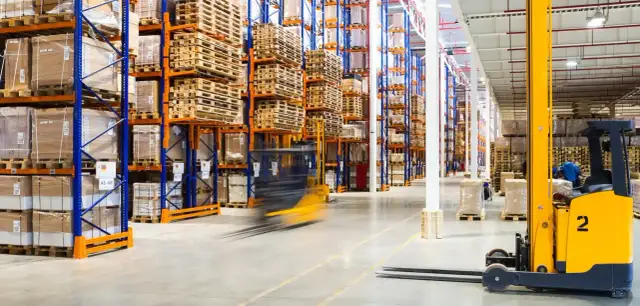 Wózek widłowy w ruchu w magazynie z wysokimi regałami pełnymi towarów i palet.