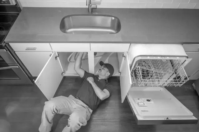 Mężczyzna pod zlewem sprawdza, jak sprawdzić elektrozawór w zmywarce, która jest otwarta.