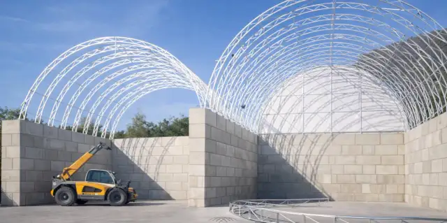 Budowa hali z pustaków szalunkowych, z widocznym zbrojeniem i żółtym ładowarką teleskopową.