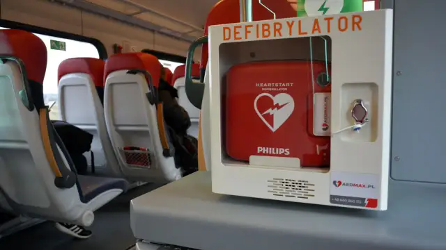 Czerwony defibrylator AED w białej obudowie, zamontowany w pociągu, obok siedzeń.