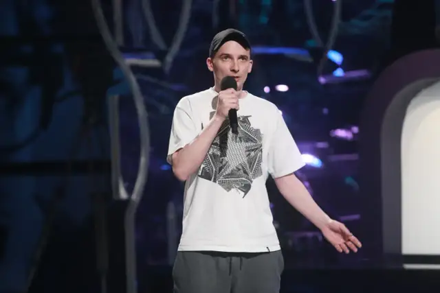 Jasiek Borkowski w czapce i białej koszulce z grafiką, trzymający mikrofon, podczas występu stand-up.