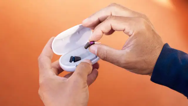 Dłonie trzymają aparat słuchowy i jego etui na pomarańczowym tle.