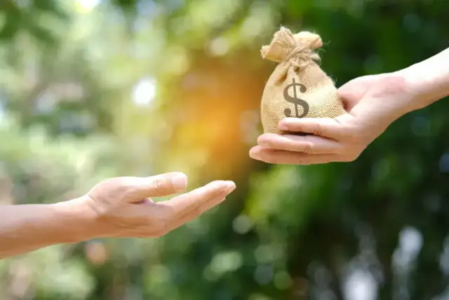 Dwie dłonie, jedna podaje woreczek z pieniędzmi z symbolem dolara, druga wyciągnięta do odbioru.