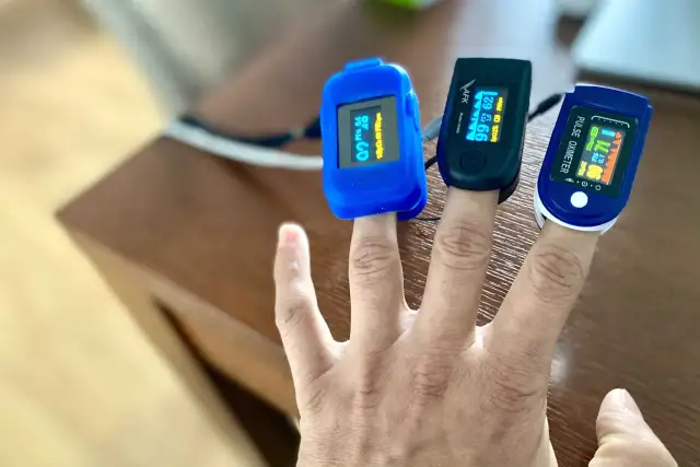 Trzy pulsoksymetry na palcach pokazują prawidłową saturację u dziecka: 99%, 99% i 99%.